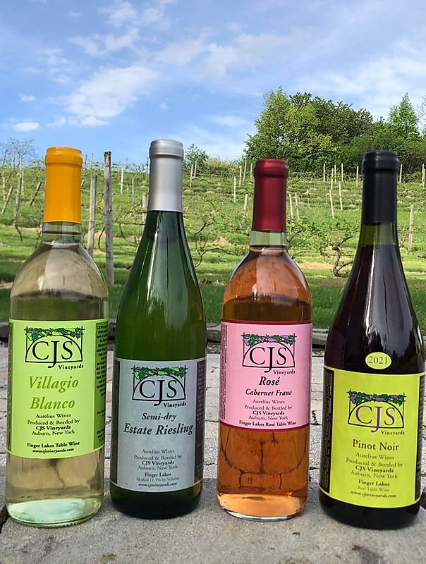 Four bottles of wine from CJS Vineyards, with vineyards in the background. The labels include Villagio Blanco, Semi-dry Estate Riesling, Rosé Cabernet Franc, and Pinor Noir.
