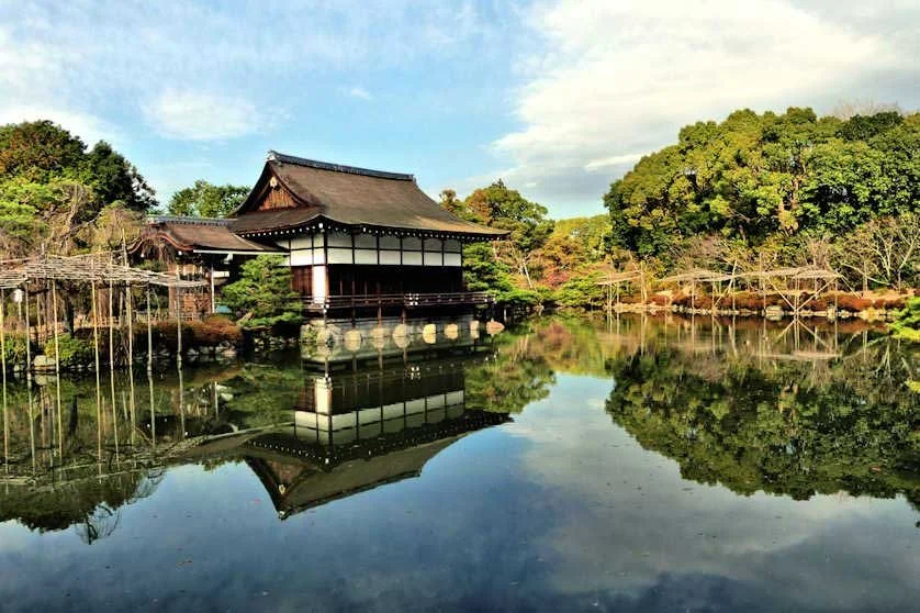 Traditional Japanese building near a calm pond with trees and sky reflected in the water.