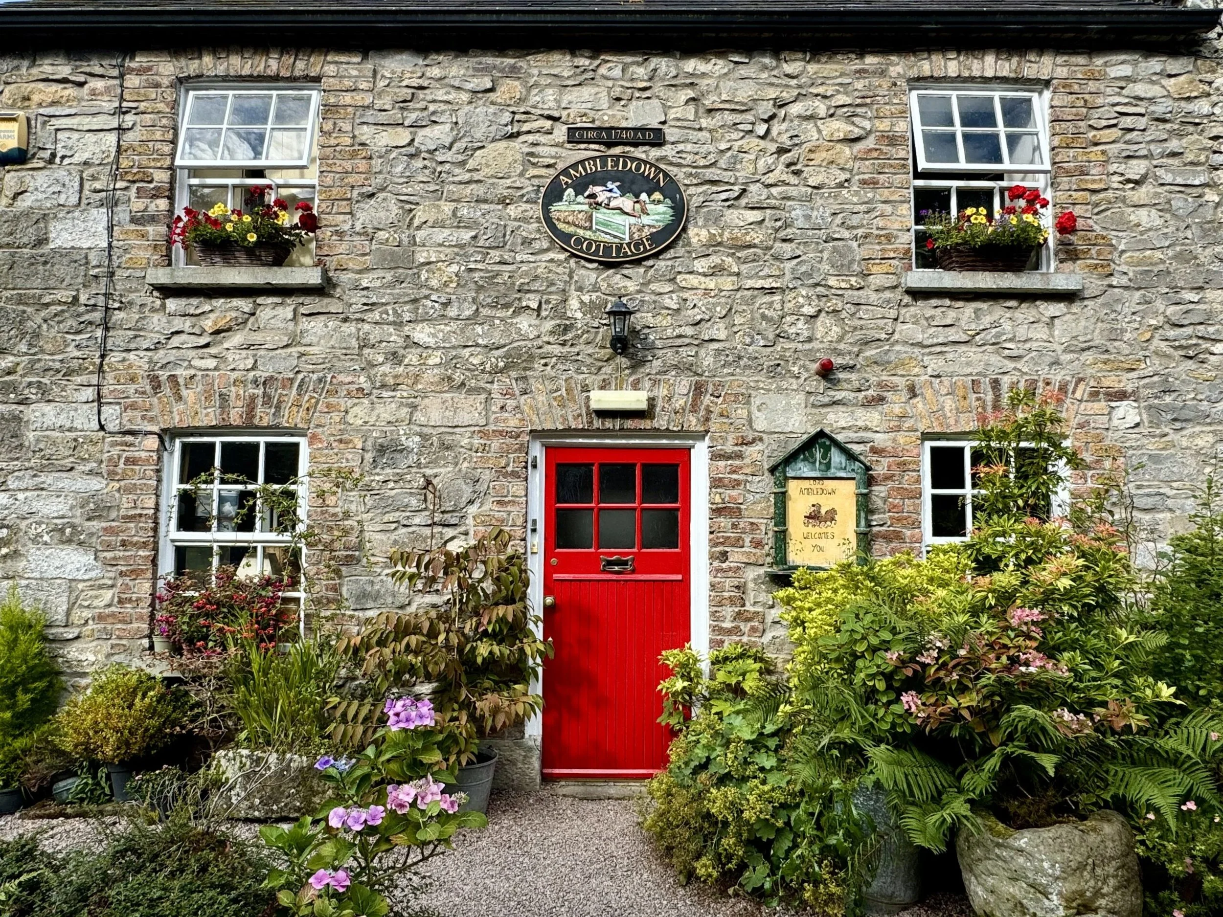 A stone cottage with two small windows and a red door, surrounded by colorful flowers and green plants, with a sign reading "Ambledown Cottage" on the wall.