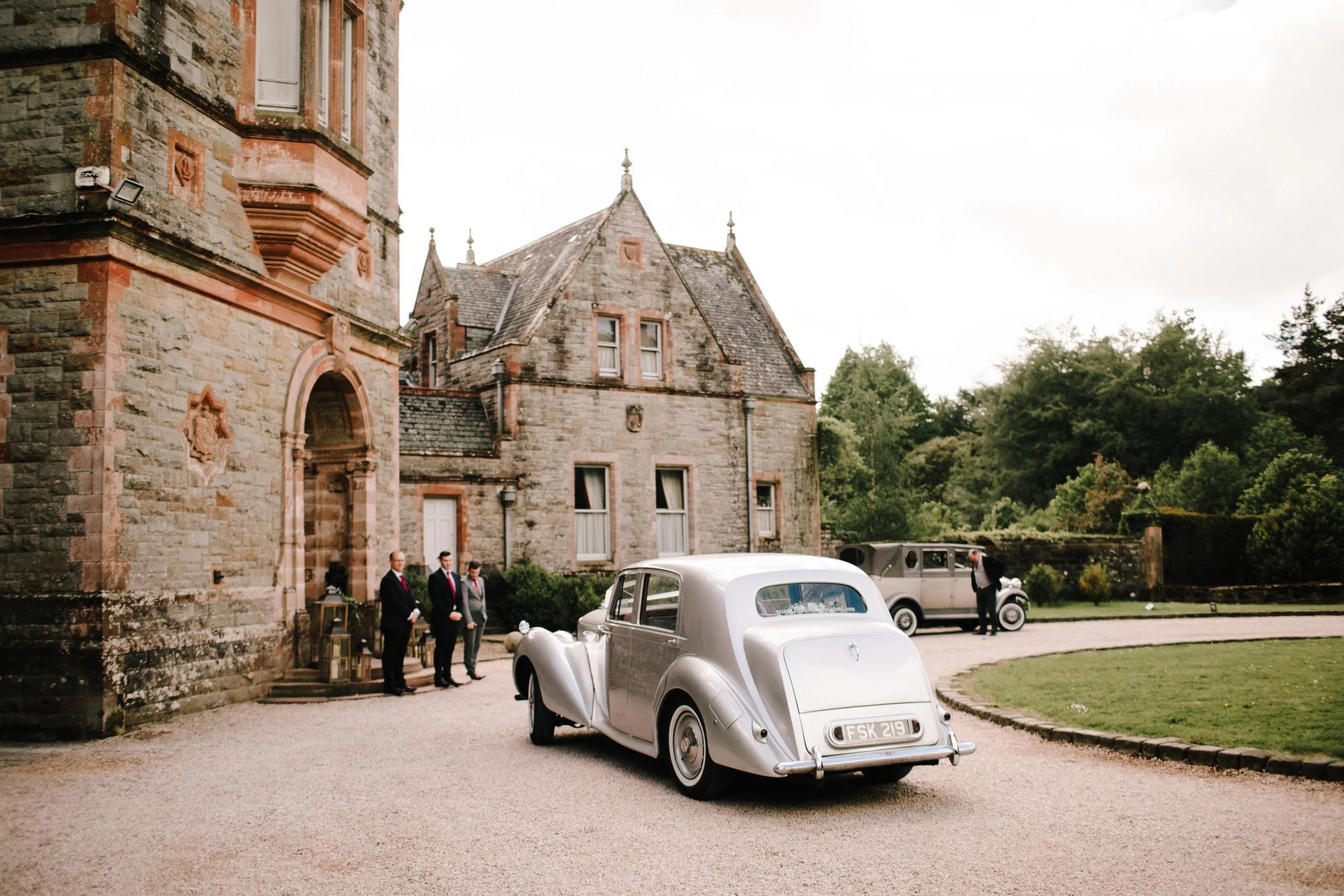 A vintage wedding scene outside a stone church with a classic white car in the foreground, four men in suits near the entrance, and a green landscape with trees in the background.