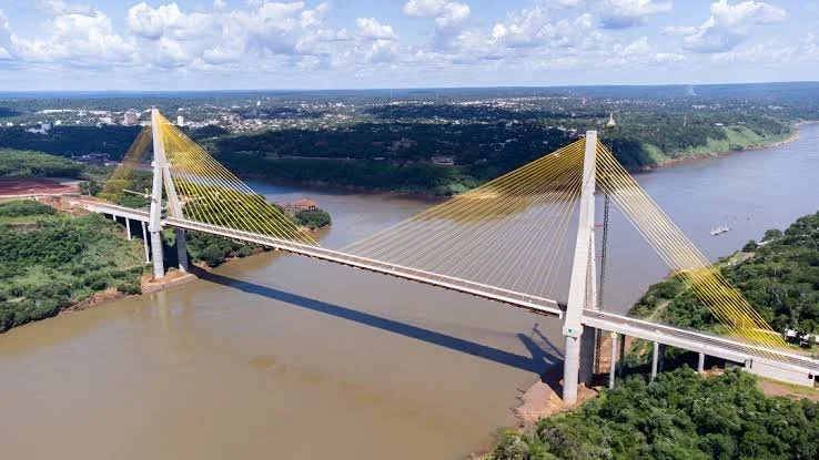 A cable-stayed bridge crossing a wide river with green landscape on both sides and a city skyline in the distance.