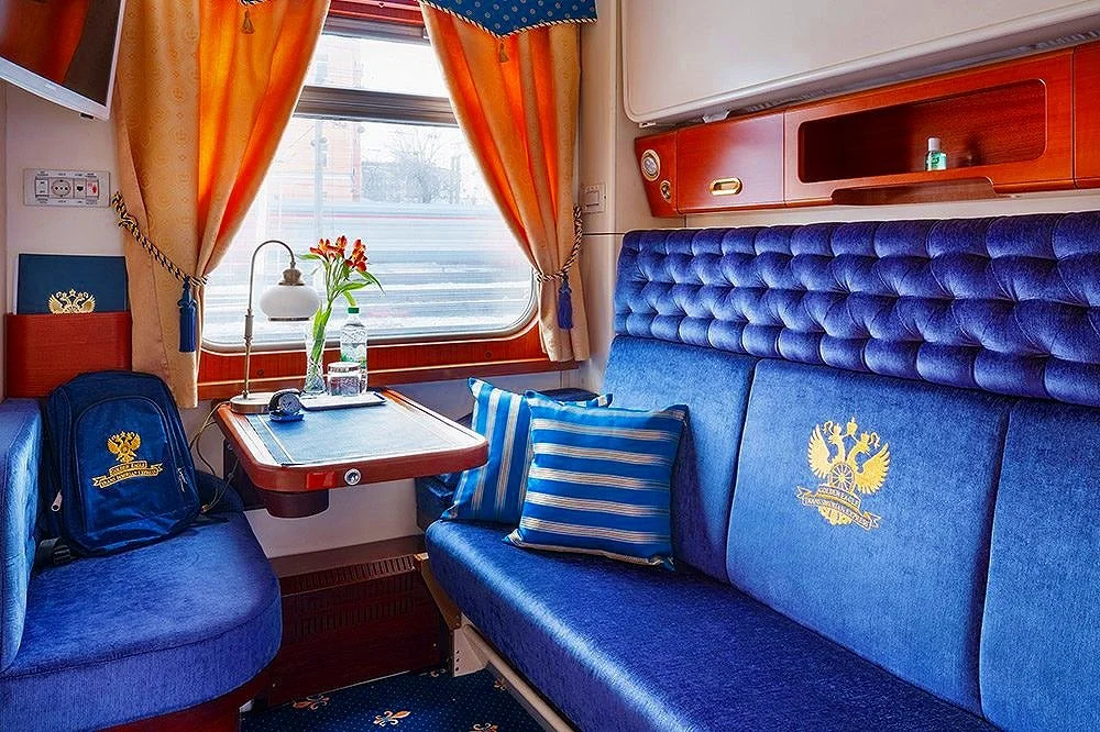 Interior view of a train compartment with blue upholstered seating, a window with orange curtains, a small table with a flower arrangement, and a backpack with a crest logo.