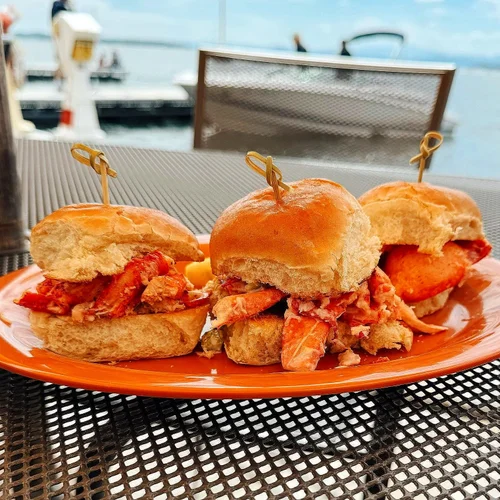 Three lobster rolls on an orange plate, with a dock and boats in the background.