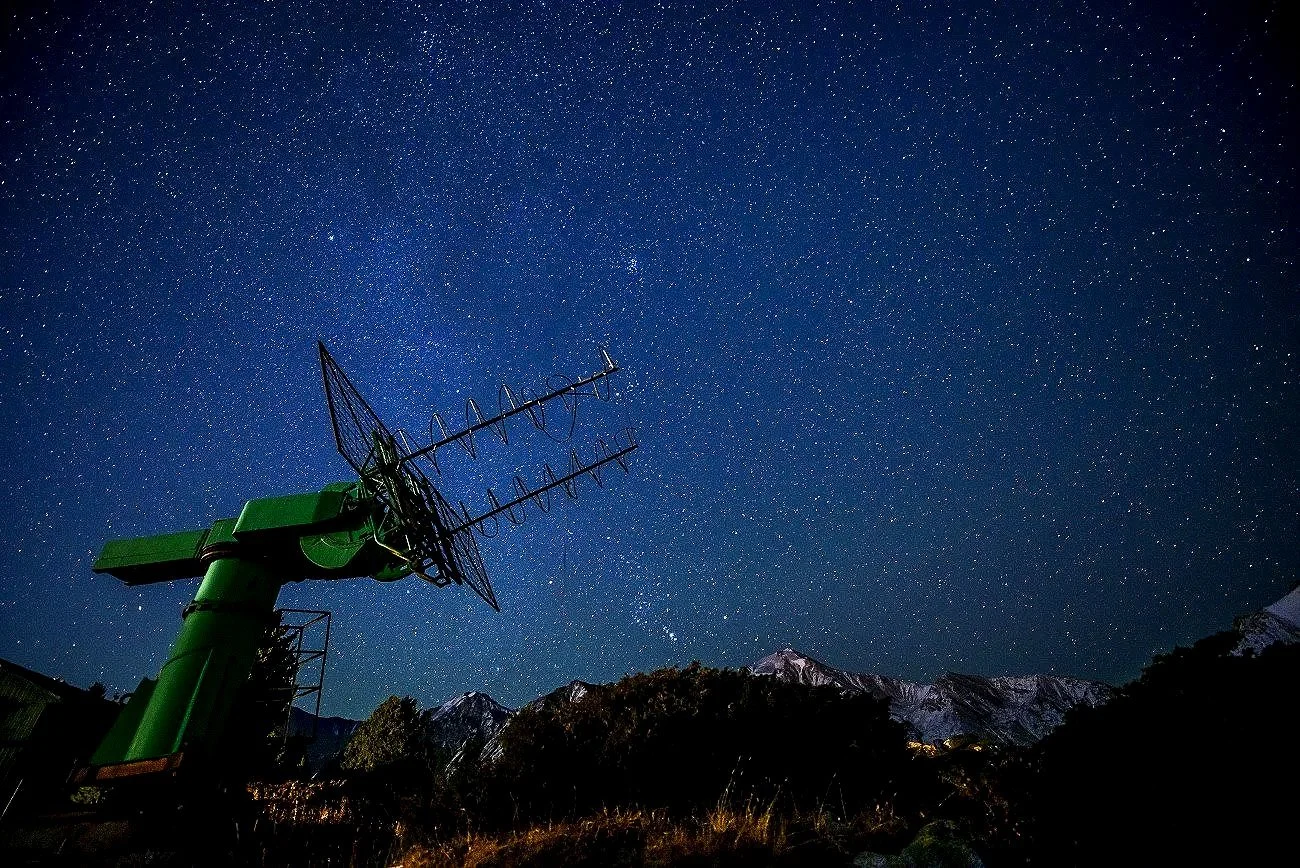 A photo of a starry night sky with a prominent mountain range in the foreground and large antenna equipment on the left side.