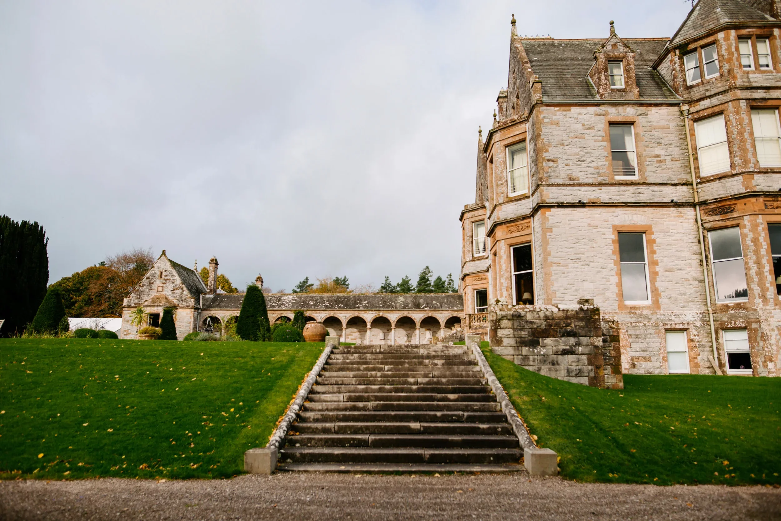 A large stone mansion with multiple windows and a steep roof, surrounded by a green lawn with stone stairs leading up to the entrance, trees, and a cloudy sky.
