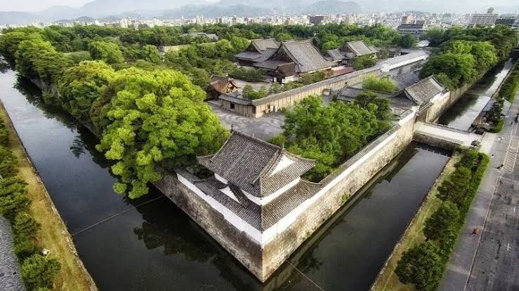 Aerial view of a traditional Asian fortress surrounded by a moat, with lush green trees and modern buildings in the background.