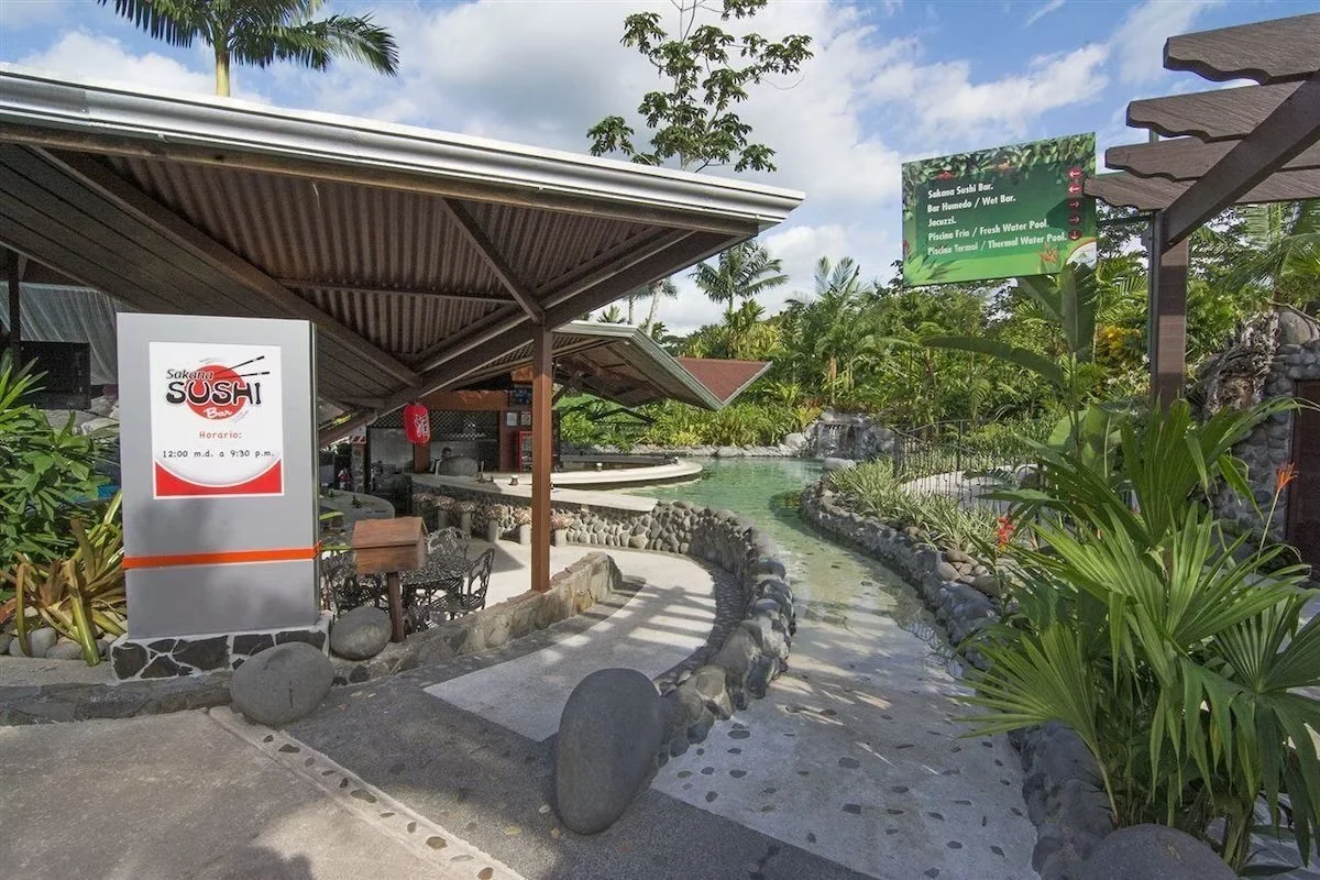 Outdoor area of a restaurant with a sign that reads "Saken SUSHI Bar" and its operating hours, surrounded by lush green tropical plants and trees, with a small stream or water feature running through the space, and a decorative stone walkway.