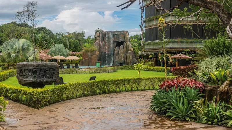 Lush tropical garden with neatly trimmed bushes, tropical plants, a waterfall feature, and a building in the background.