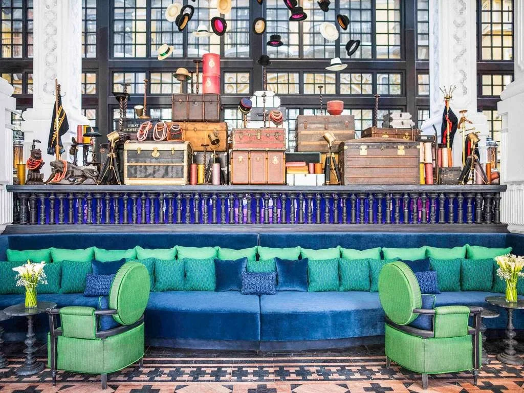 A colorful hotel lobby with a vibrant blue and green seating area in front and a large display of vintage luggage and hats above. The luggage and hats are arranged on a dark wood display shelf with a black railing, and the background features large w