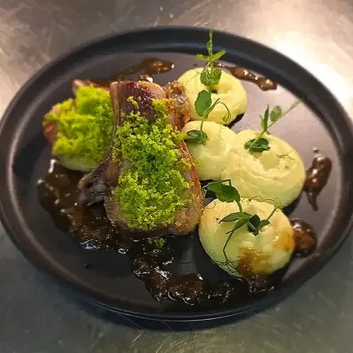 A plated dish with a meat chop, green puree, four scoops of white mashed potatoes garnished with microgreens, and dark sauce on a black plate.