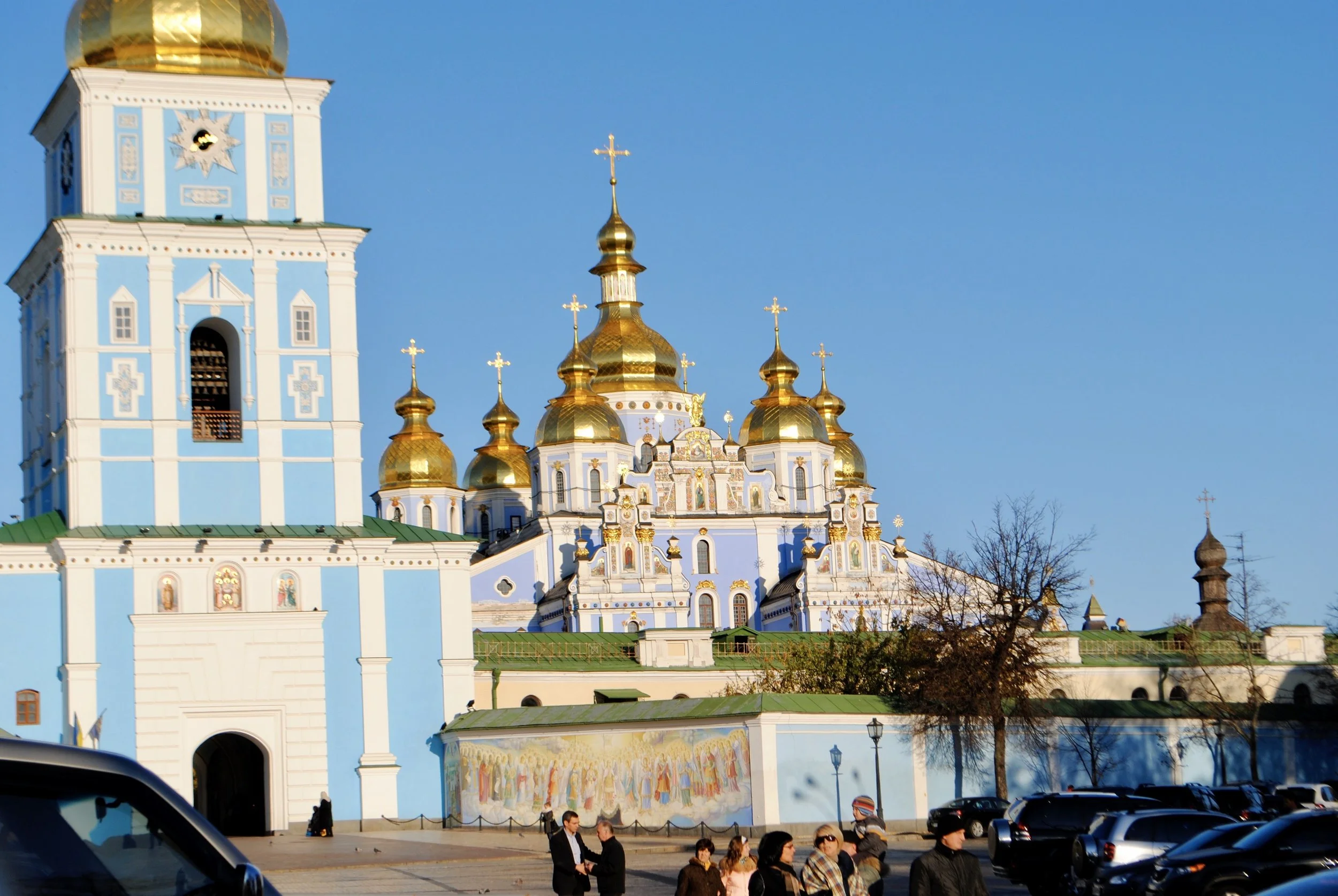 People walking near a church with golden domes and a tall clock tower, under a clear blue sky.