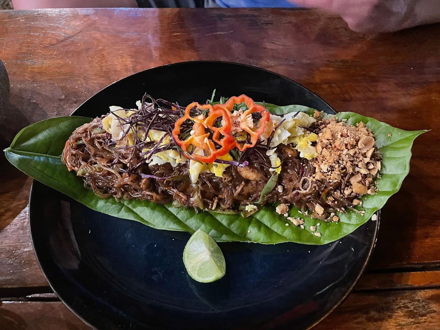 Plate of stir-fried noodles with vegetables, topped with sliced red bell peppers and shredded purple cabbage, served on a large leaf with lime wedge on a black plate.