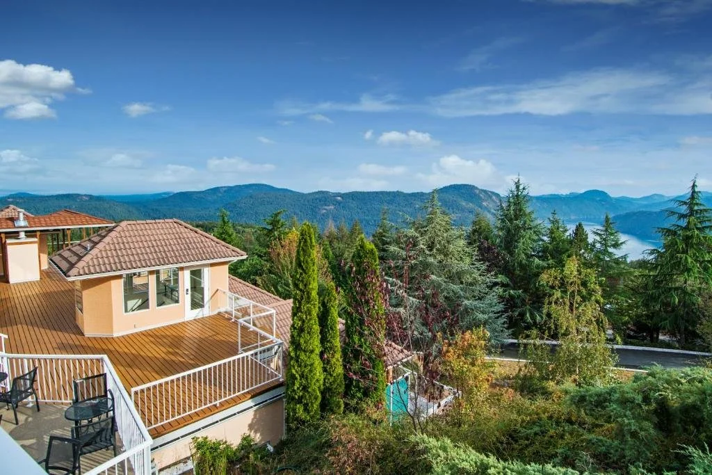 A view of a house with a large wooden deck, surrounded by tall trees and lush green mountains in the background under a partly cloudy sky.