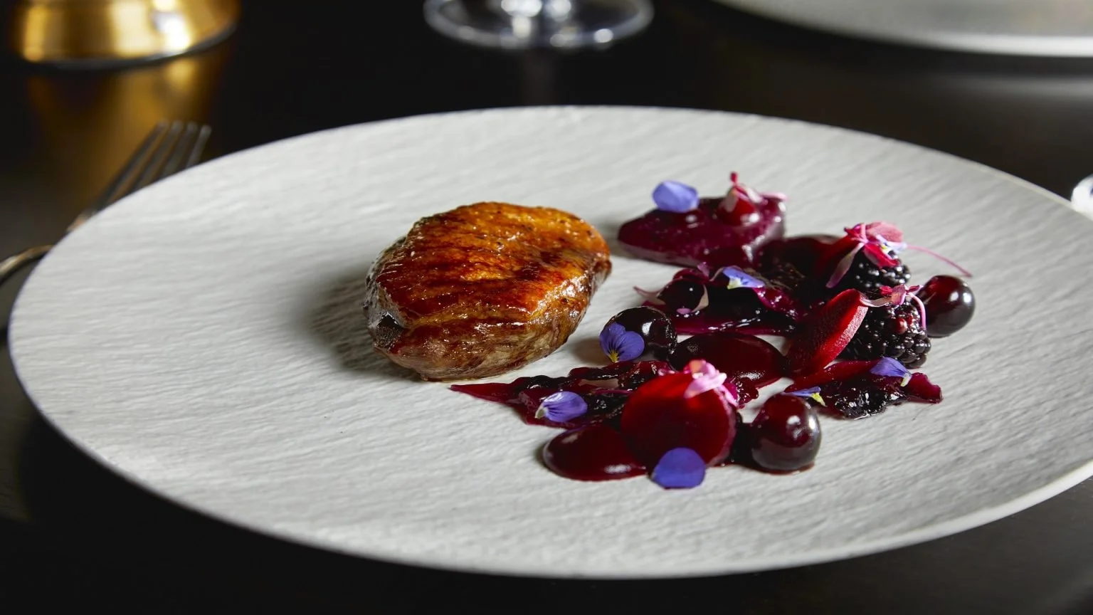 Seared meat served with a variety of berry compote and edible purple flowers on a white textured plate.