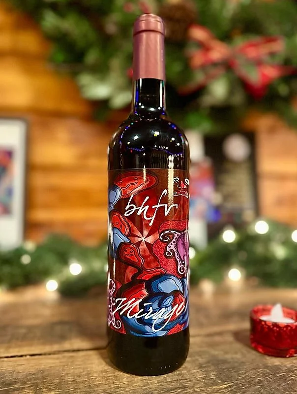 A bottle of wine with a colorful label featuring red and blue ink-like designs, the words 'bff' and 'merry' written in white script, on a rustic wooden table with holiday decorations blurred in the background.