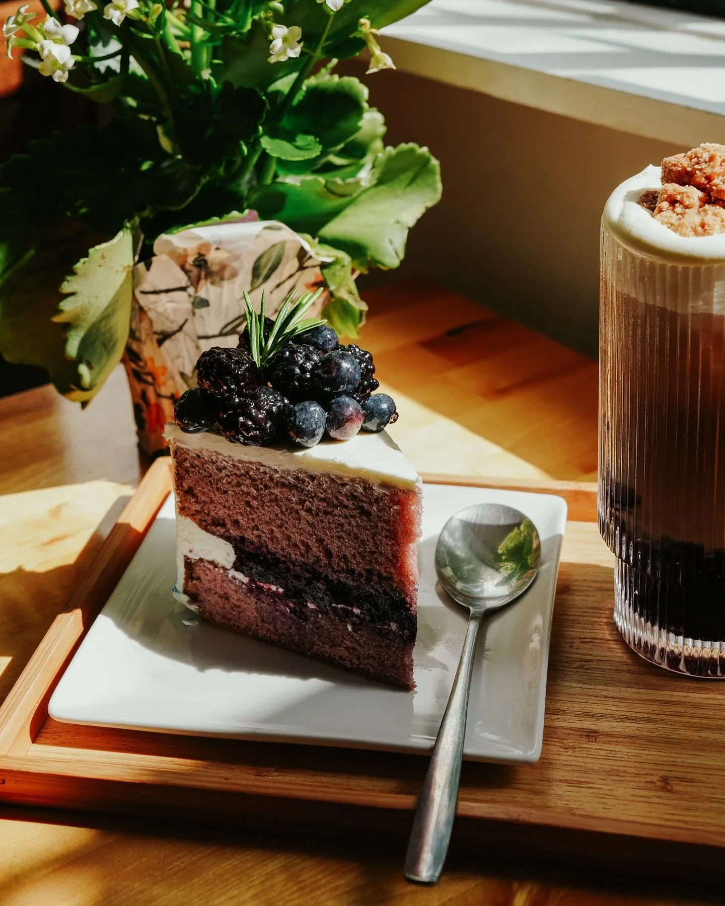 A slice of berry cake topped with blackberries and blueberries, a sprig of rosemary, with a spoon on a white plate, alongside a tall glass of soda with whipped cream, on a wooden table with green plants in the background.