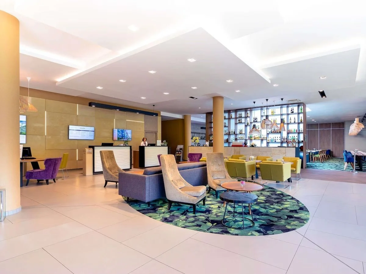 Lobby of a hotel or lounge with seating areas, a bar shelf in the background, and a reception desk with a staff member behind it.