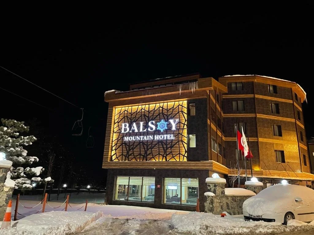 A multi-story hotel building at night with a lit sign that reads "BALS Y MOUNTAIN HOTEL," with snow on the ground and over the car parked outside, and ski chairs hanging above.