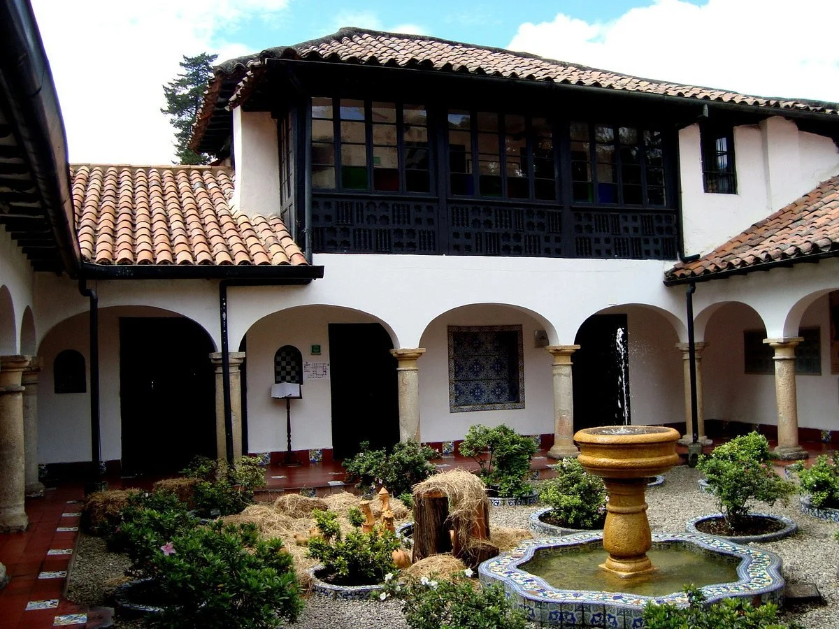 Interior courtyard of a Spanish-style building with a central fountain, balcony with black wooden latticework, arched walkways, and lush small bushes and plants.