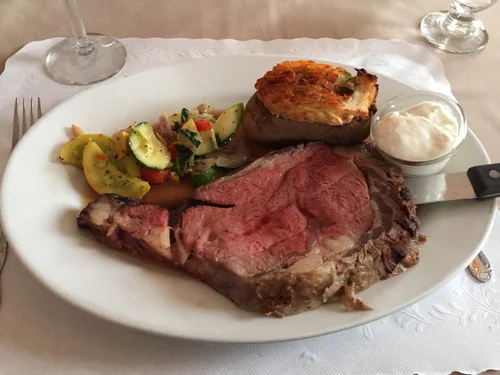 Plate with cooked prime rib, baked potato topped with cheese and bacon, mixed vegetable salad, and a bowl of creamy horseradish sauce, on a white tablecloth with silverware and wine glasses.