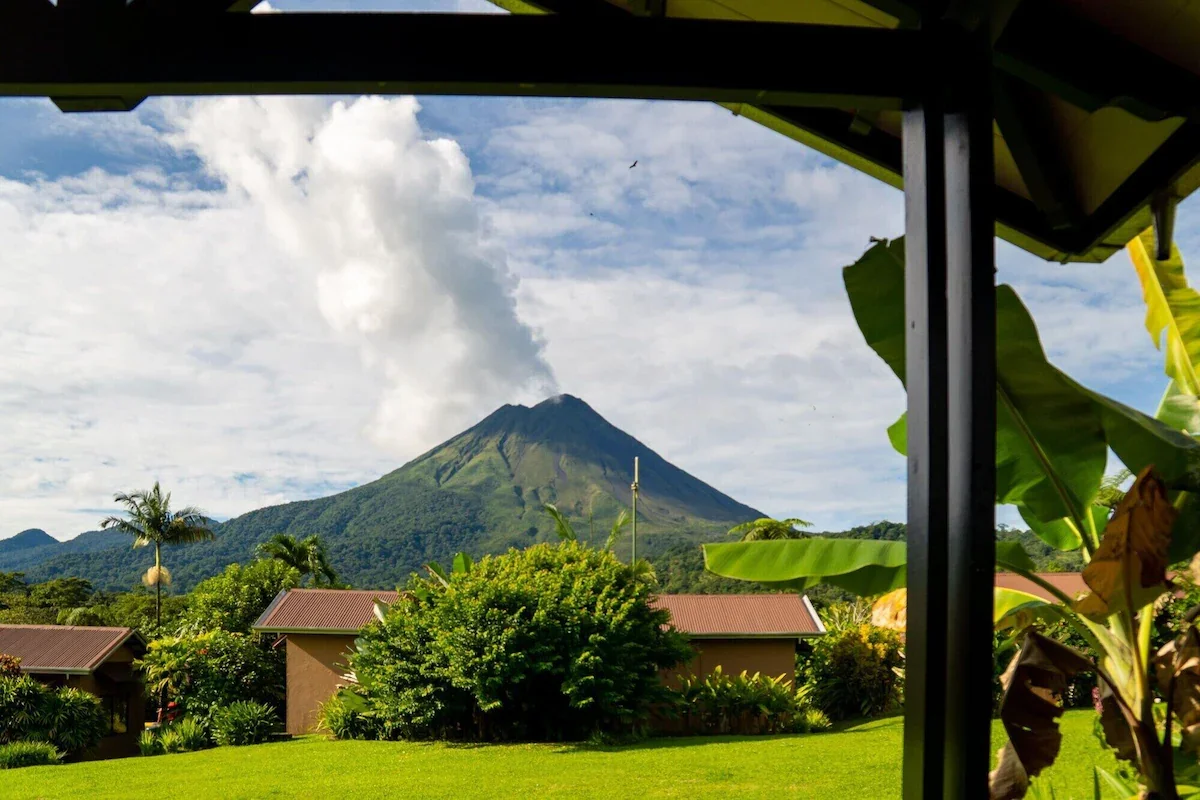 View of a smoking volcano with lush green landscape and houses in the foreground, seen through a covered area with plants.