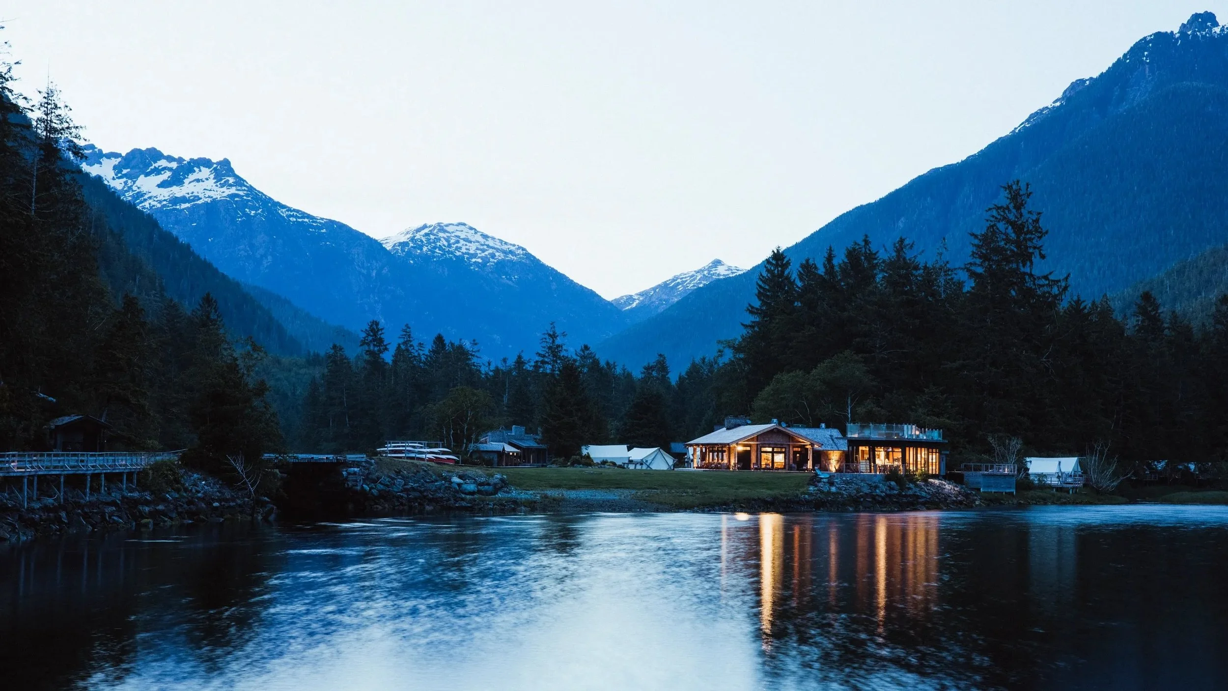 A lakeside house with lights on, nestled among trees with snow-capped mountains in the background, at dusk.