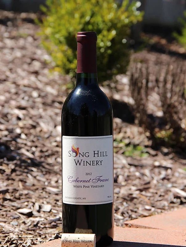 A bottle of 2012 Cabernet Franc wine from Song Hill Winery, standing upright outdoors on a brick surface with a blurred background of mulch, green bushes, and sunlight.