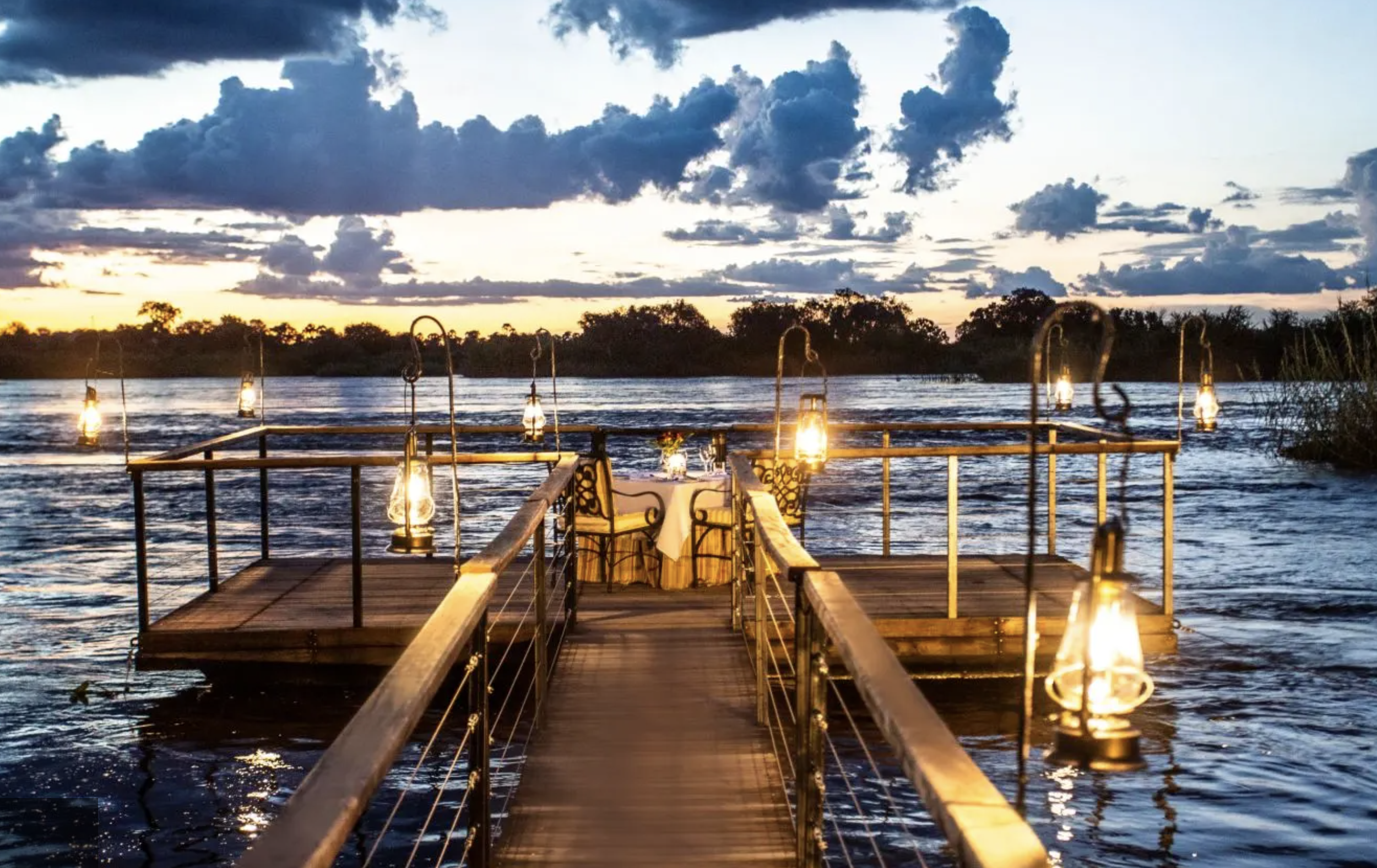 A wooden dock with a table set for dining, adorned with lanterns, extends into a river at sunset, with clouds in the sky and trees on the horizon.