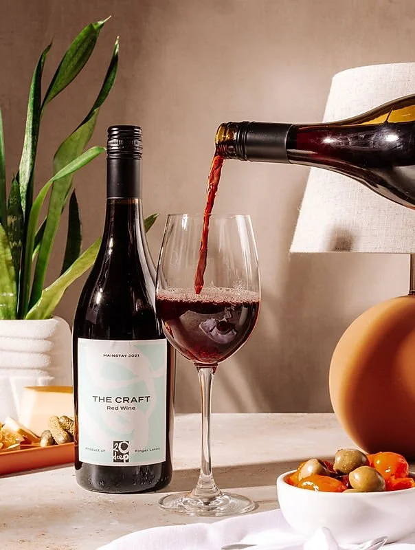 A bottle of red wine labeled 'The Craft' is being poured into a glass, with another bottle labeled 'Mainstay 2021' nearby. There is a potted plant to the left and a bowl of colorful olives to the right, all on a light-colored surface in a cozy indoor setting.