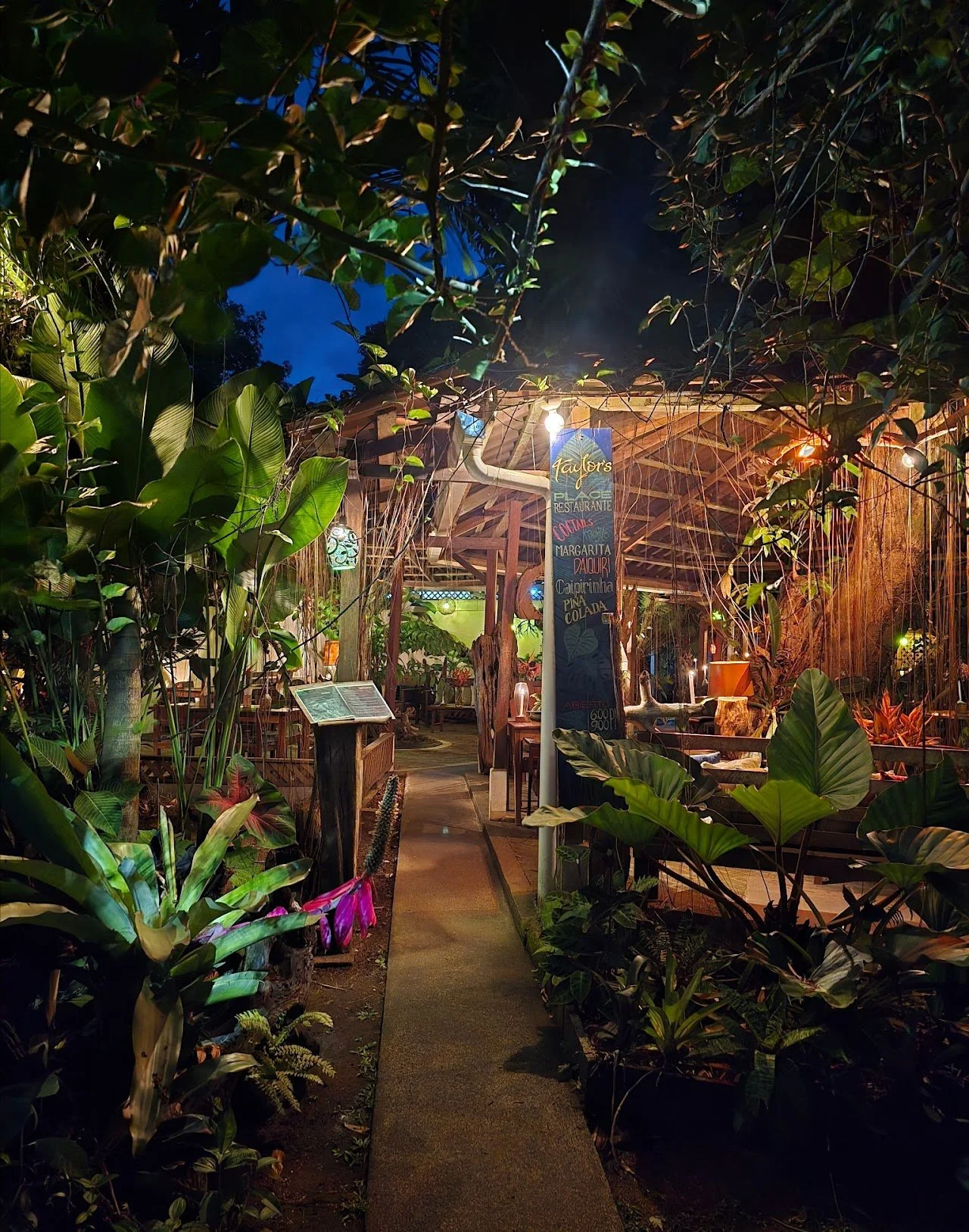A cozy outdoor restaurant or bar area illuminated with warm lighting, surrounded by lush green tropical plants and trees, with a narrow pathway leading to the seating area and a menu stand on the side.