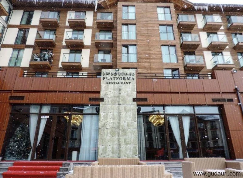 Side view of a modern multi-story building with balconies, a sign with Georgian script and English words that reads "Plattform Restaurant," and snow falling in front of the building.