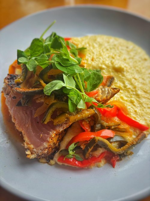 A plate with a serving of baked fish topped with sautéed peppers and greens, served with a creamy side of polenta.