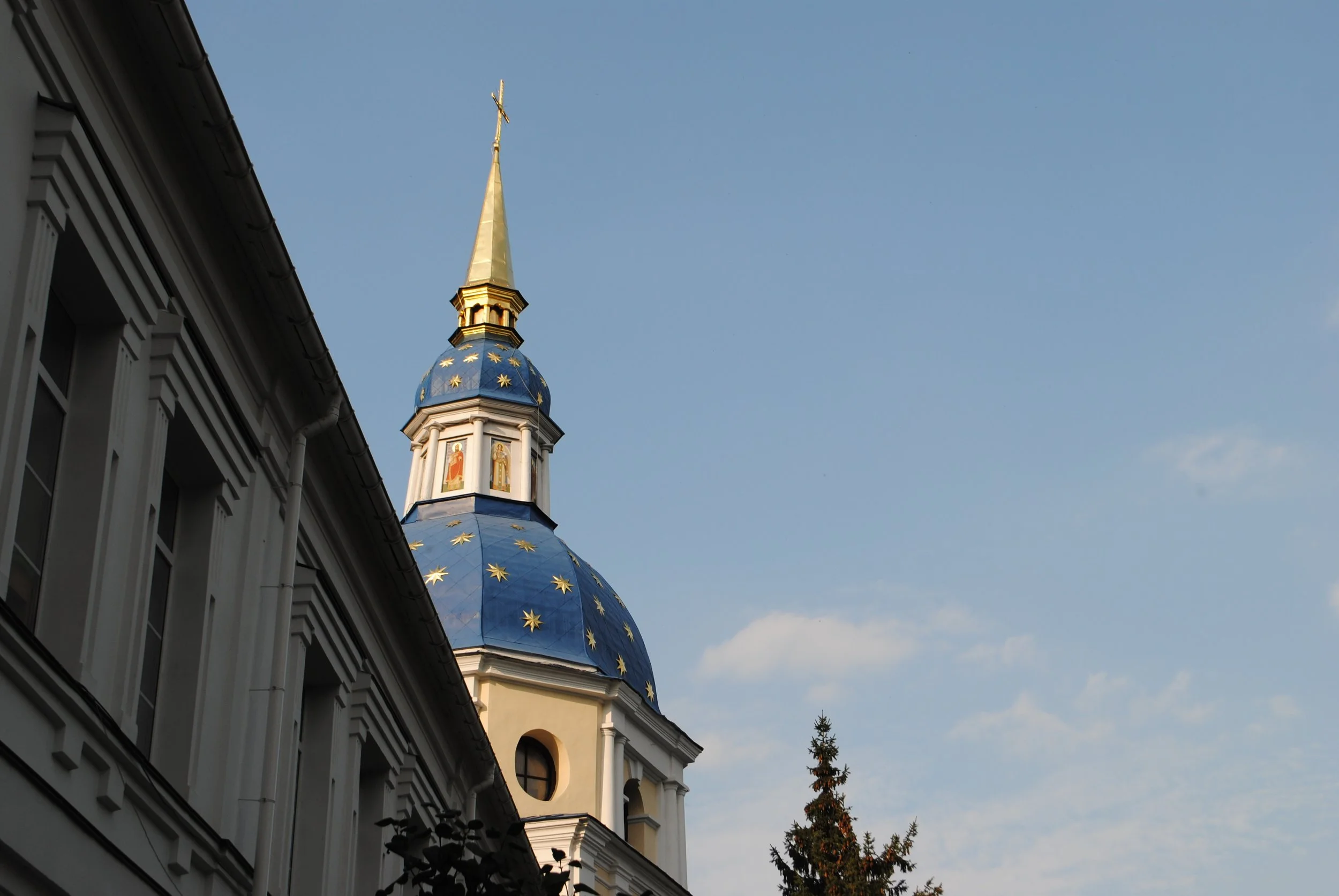 View of a church steeple with a golden cross, blue roof decorated with stars, and part of the church building visible on the left, against a clear blue sky.