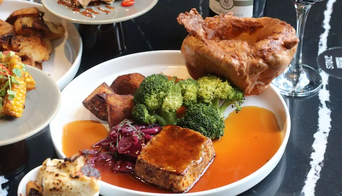 A roasted pork shank with crispy skin on a white plate, served with broccoli, a meatloaf slice, and red cabbage, with gravy.