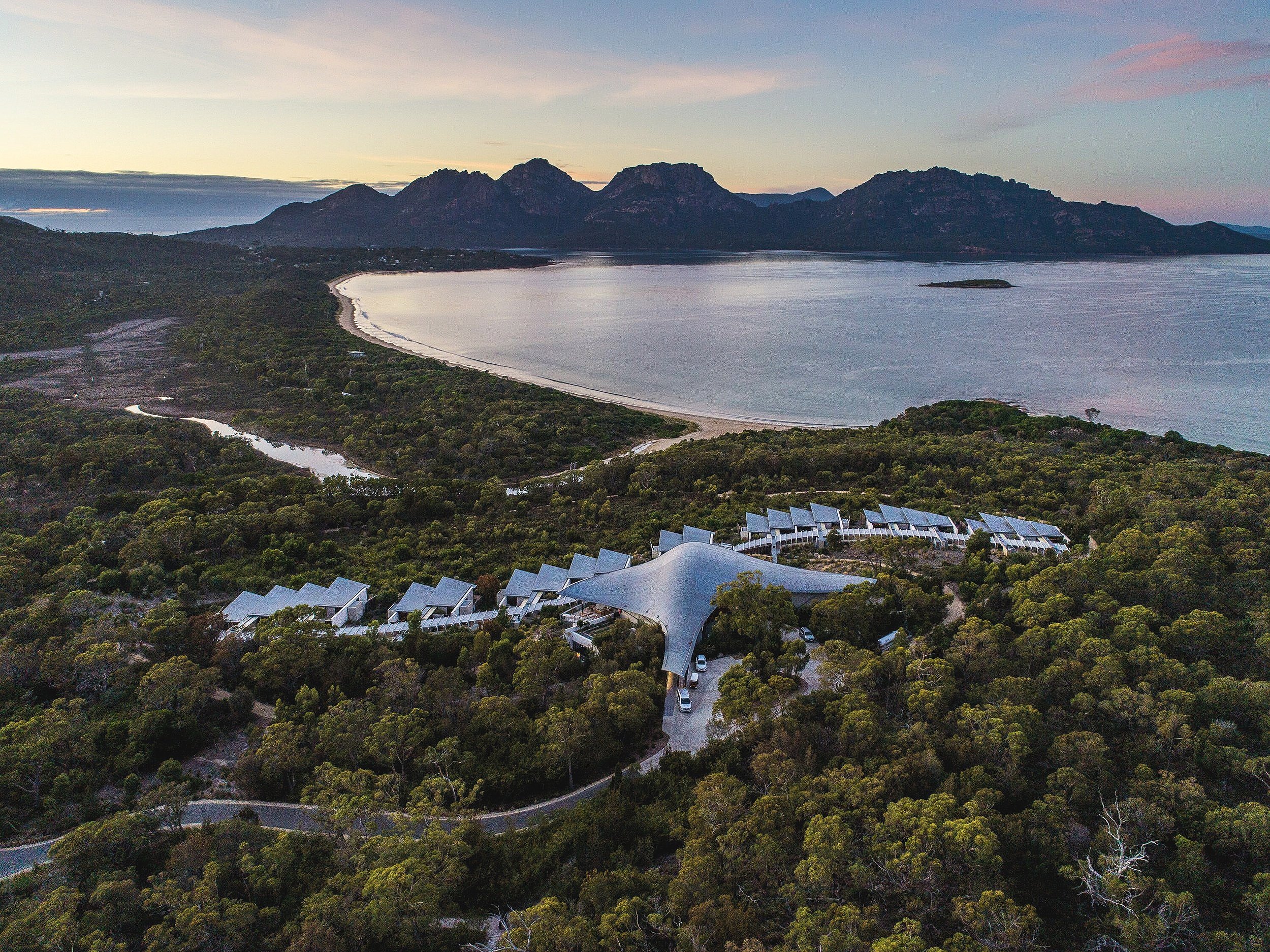 Aerial view of modern resort buildings with a large canopy roof surrounded by forest, near a coastal bay at sunset, with mountains in the background.