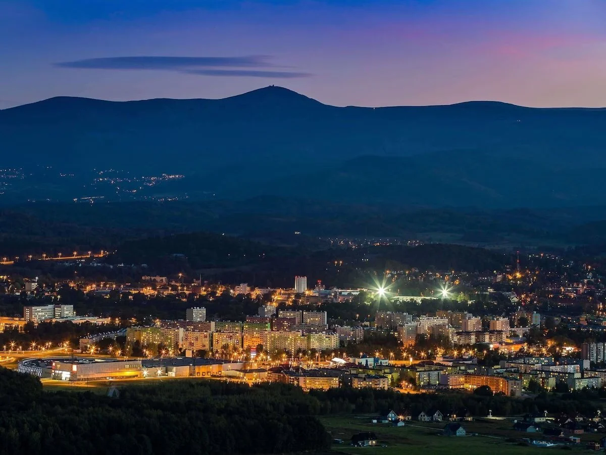 Nighttime cityscape with brightly lit buildings and streetlights, mountains in the background under a darkening sky.