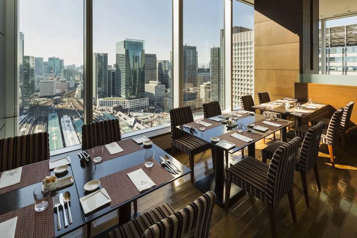 A modern restaurant with a view of a city skyline through large glass windows, featuring dining tables set with plates, glasses, and utensils