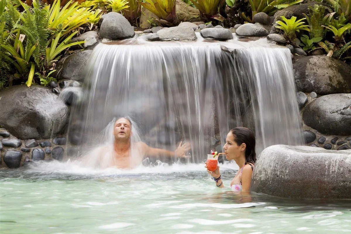 A man is relaxing under a small waterfall in a pool, with a woman nearby holding a drink with a umbrella and fruit garnish.