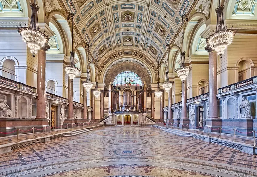 The image shows an ornate, grand hall with a high, arched ceiling decorated with intricate patterns and gold accents. There are large chandeliers hanging from the ceiling, illuminating the space. The hall features marble columns and statues along the