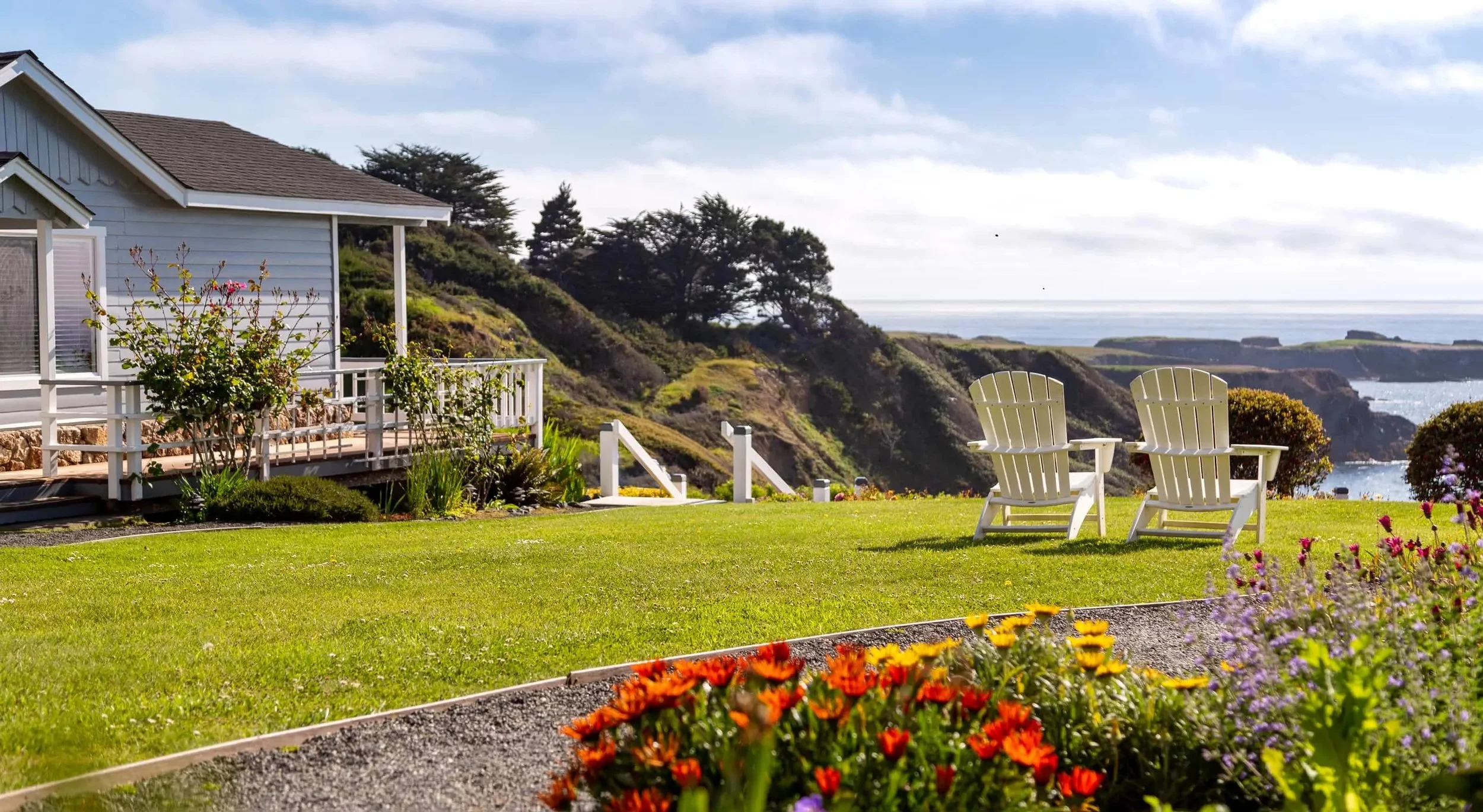 A backyard with two white Adirondack chairs facing the ocean, surrounded by colorful flowers, green lawn, and a house with a deck, overlooking rocky cliffs by the sea.