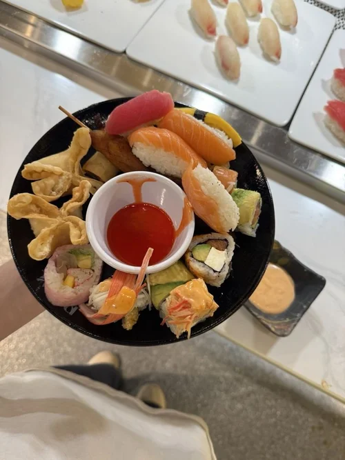 A hand holding a black plate with assorted sushi, tempura, and a small bowl of soy sauce in the center. In the background, there are more sushi pieces on white plates.