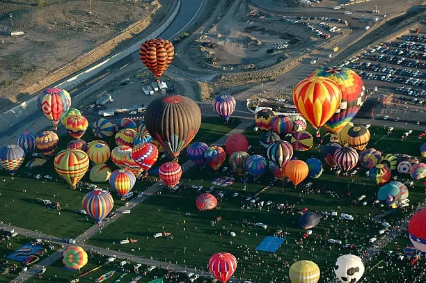 Aerial view of a hot air balloon festival with numerous colorful hot air balloons in various shapes and patterns, some near the ground and others ascending into the sky.