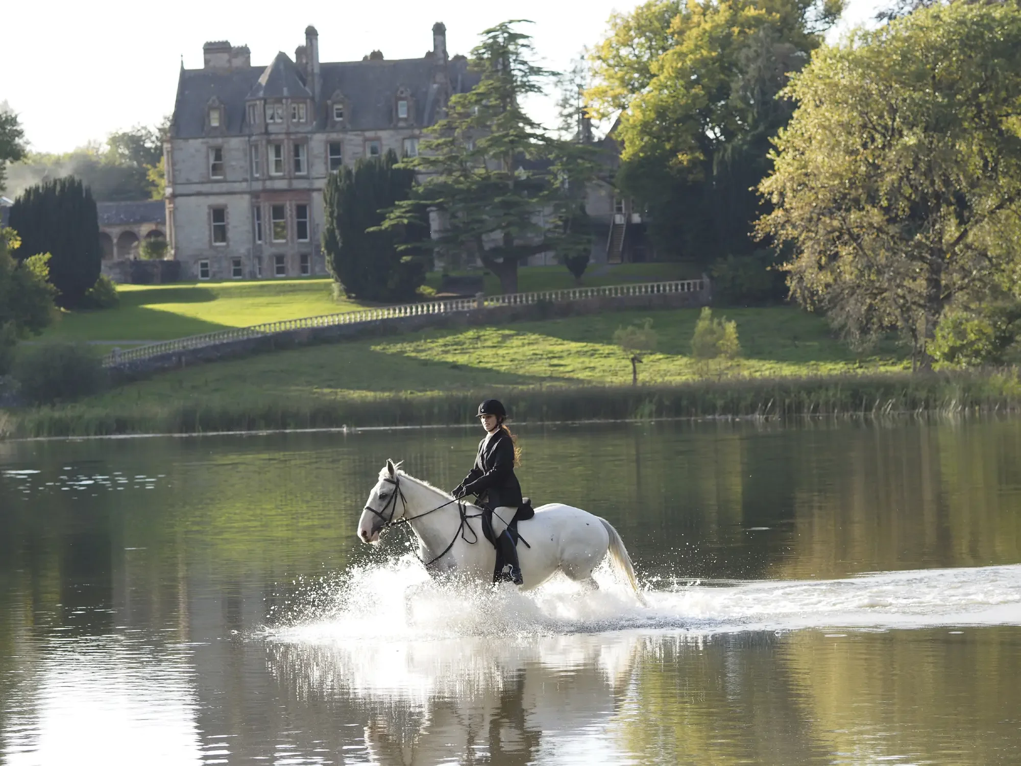 A person riding a white horse through a body of water with a large castle and trees in the background.