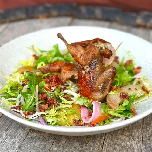 Bowl of fresh salad with grilled chicken pieces, mixed greens, shredded lettuce, red onion, and bacon bits on a wooden surface.