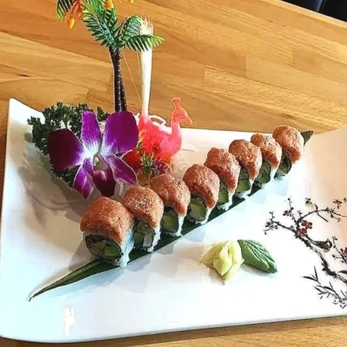A decorative platter featuring sushi rolls arranged to resemble a boat, with colorful edible decorations including purple orchid, pink flamingo, and palm tree. The platter has wasabi and ginger on the side, and an artistic floral design on the plate.
