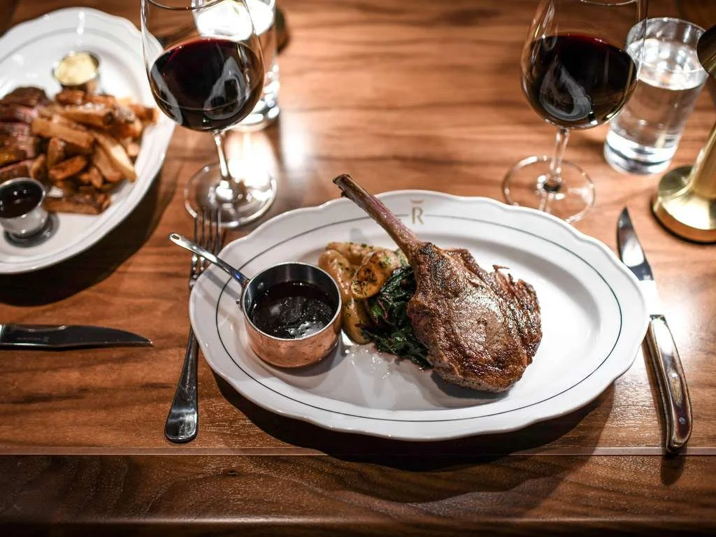 A fancy dinner plate with a lamb chop, roasted potatoes, and greens on a white plate, with a small copper cup of sauce. Two glasses of red wine and a glass of water are on the table, along with silverware.