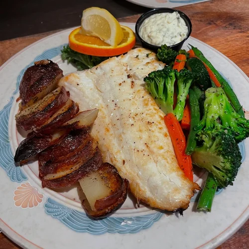 A plate with baked fish fillet, roasted potatoes, grilled vegetables including broccoli, carrots, and zucchini, lemon wedges, and a side of tartar sauce.
