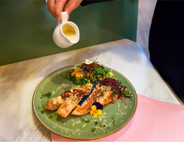 Person pouring a sauce or dressing over a plated dish that includes a cooked lobster tail, salad greens, and edible flowers on a green plate.