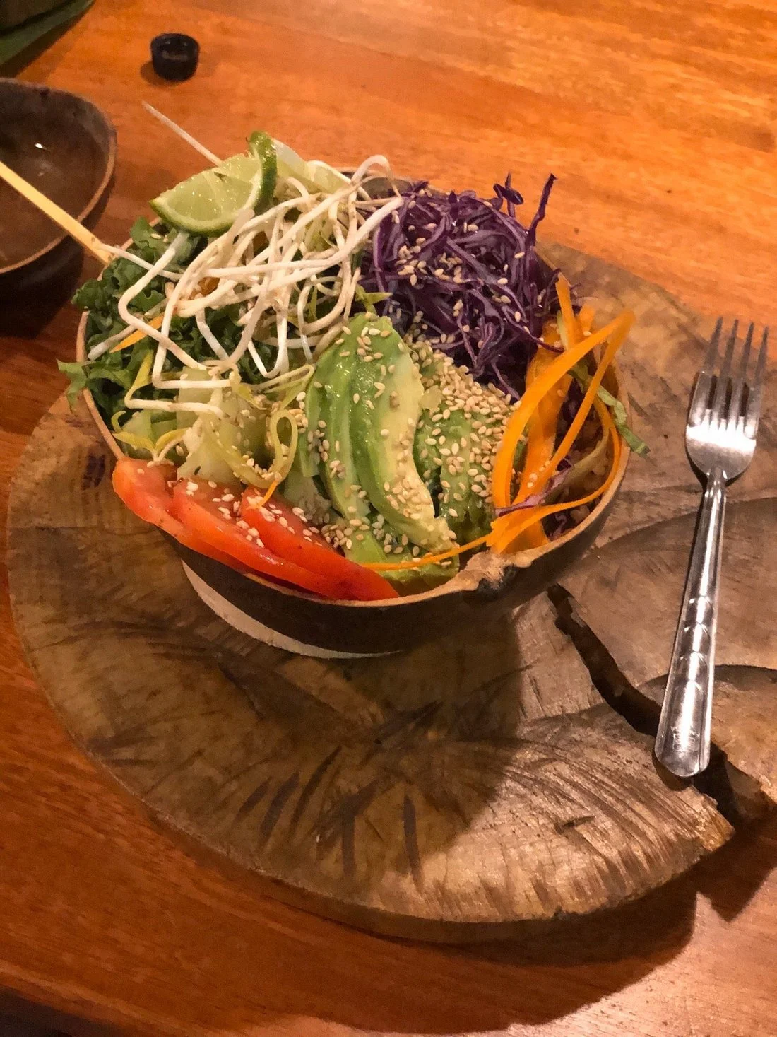 A colorful salad with lettuce, avocado, shredded carrots, purple cabbage, bean sprouts, lime wedges, tomato slices, and sesame seeds in a bowl on a wooden serving board, with a fork beside it.