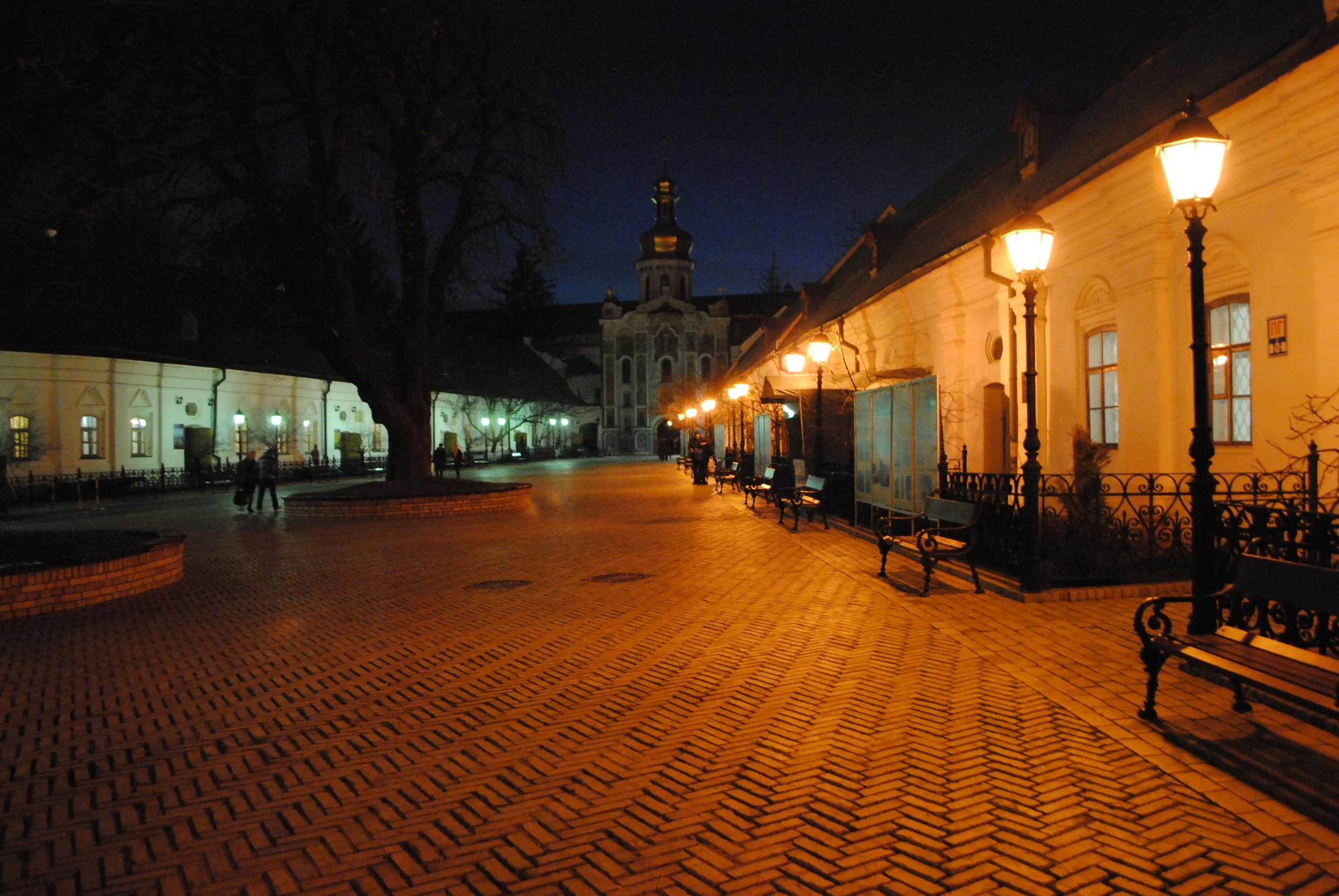 The image shows a quiet city street at night, illuminated by street lamps. There are a few people walking and sitting on benches. On the right, there are historic buildings with lit windows and ornate architecture. In the background, a church steeple